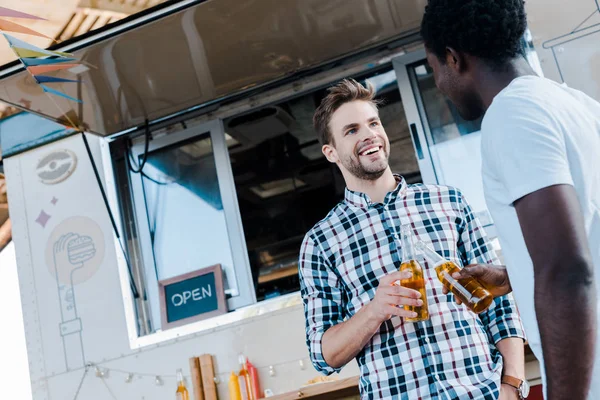 Baixo ângulo de visão de amigos multiculturais sorridentes brindar garrafas de cerveja perto de caminhão de alimentos — Fotografia de Stock
