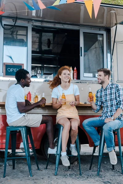 Beaux hommes multiculturels assis et tenant des bouteilles de bière près de femme rousse attrayante et camion de nourriture — Photo de stock