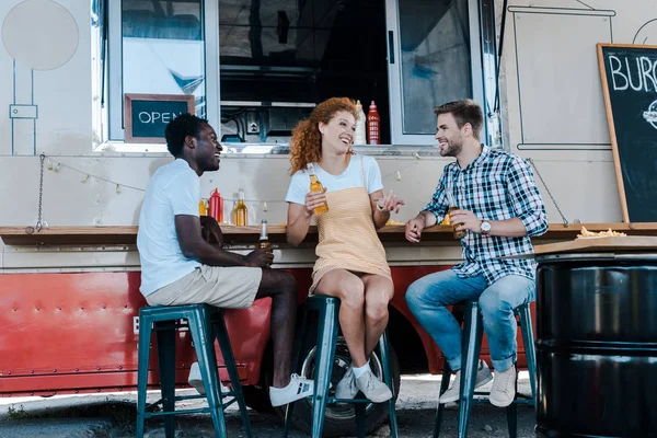 Allegri uomini multiculturali seduti e in possesso di bottiglie di birra vicino attraente donna rossa e camion cibo — Foto stock