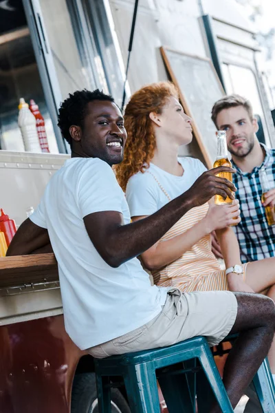 Selektiver Fokus eines glücklichen afrikanisch-amerikanischen Mannes mit einer Flasche Bier in der Nähe von Freunden — Stockfoto