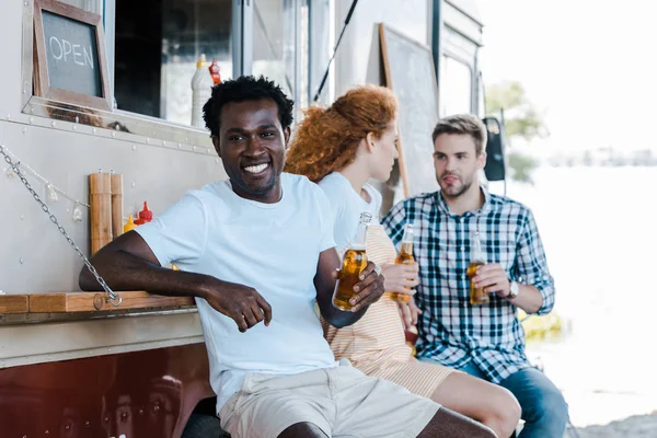 Messa a fuoco selettiva di allegro uomo afroamericano in possesso di una bottiglia di birra vicino agli amici — Foto stock