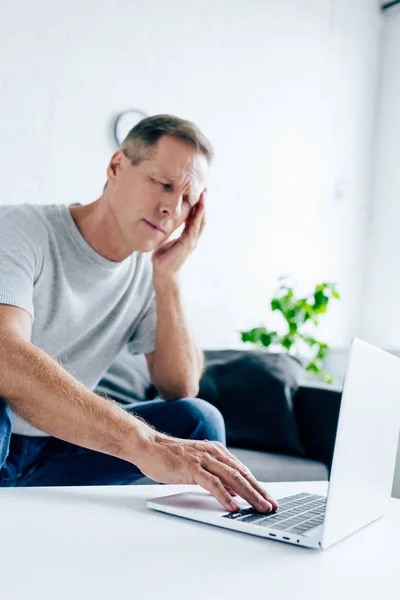 Bellissimo uomo in t-shirt con mal di testa utilizzando il computer portatile in appartamento — Foto stock