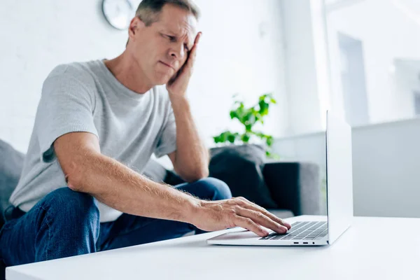 Bellissimo uomo in t-shirt con mal di testa utilizzando il computer portatile in appartamento — Foto stock