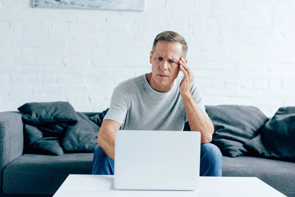 Hombre guapo en camiseta con dolor de cabeza usando el ordenador portátil en el apartamento - foto de stock