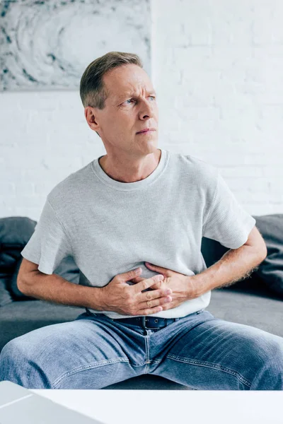 Hombre guapo en camiseta con dolor de estómago mirando hacia otro lado en el apartamento - foto de stock