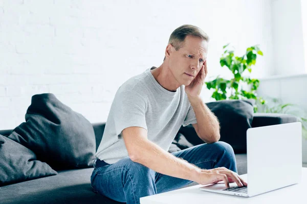 Homem bonito em t-shirt com dor de cabeça usando laptop no apartamento — Fotografia de Stock