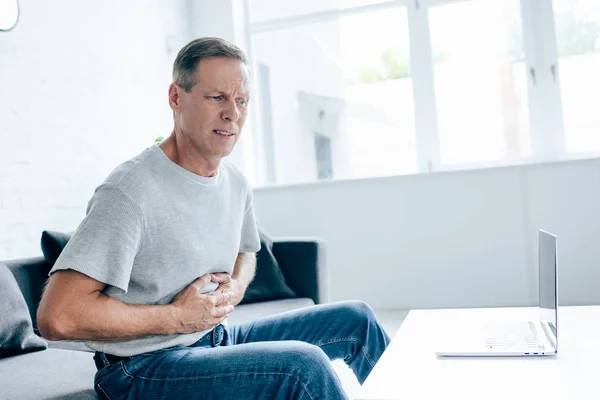 Handsome man in t-shirt with stomachache sitting on sofa — Stock Photo