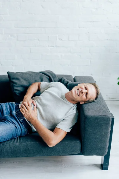 Красивий чоловік у футболці з болем в животі лежить на дивані — стокове фото