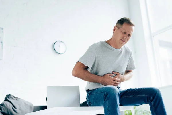 Handsome man in t-shirt with stomachache sitting on table — Stock Photo