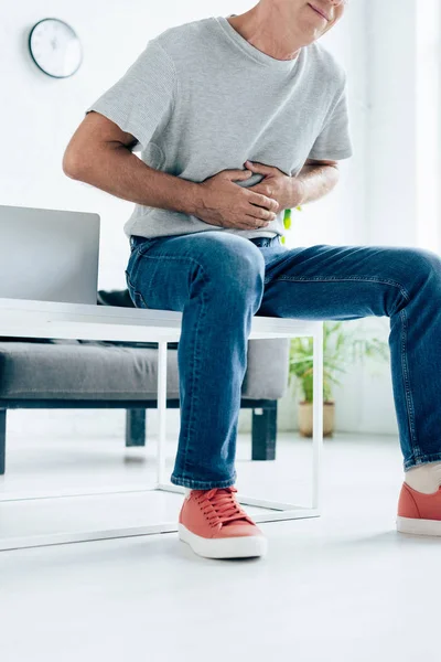 Обрезанный вид человека в футболке с болью в животе, сидящего на столе — стоковое фото