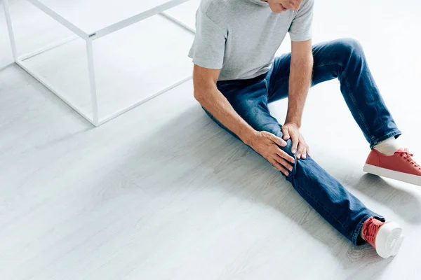 Cropped view of man in t-shirt feeling pain in knee in apartment — Stock Photo
