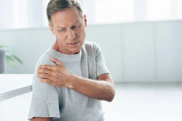 Bello uomo in t-shirt sensazione di dolore alla spalla in appartamento — Foto stock