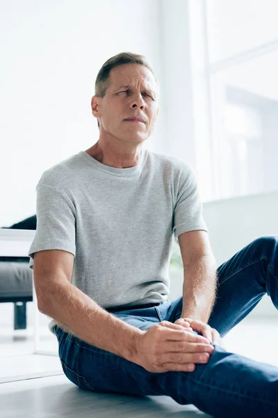 Hombre guapo en camiseta sintiendo dolor en la rodilla en el apartamento - foto de stock