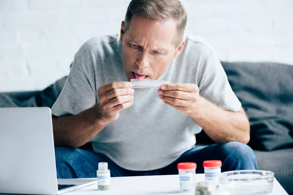 Handsome man in t-shirt licking paper for blunt with medical cannabis — Stock Photo