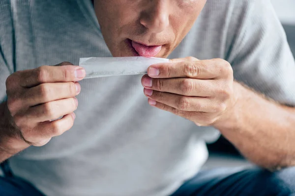 Cropped view of man in t-shirt licking paper for blunt — Stock Photo