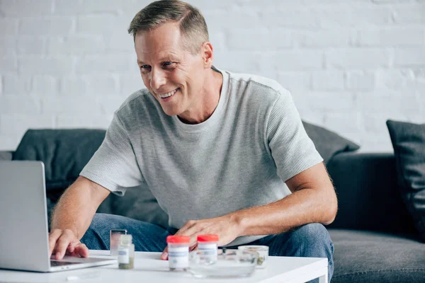 Красивый и улыбающийся мужчина в футболке с ноутбуком в квартире — стоковое фото