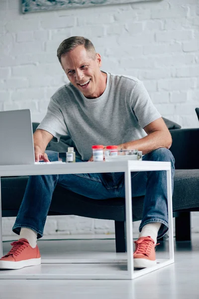 Schöner Mann im T-Shirt lächelt und benutzt Laptop in Wohnung — Stockfoto