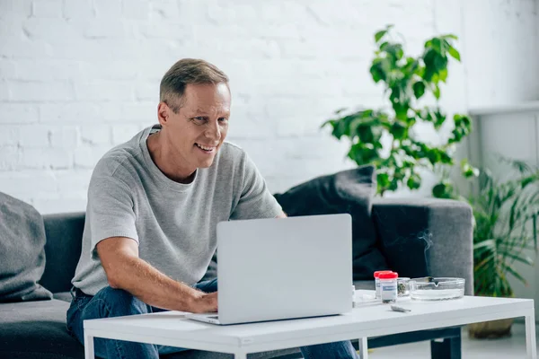 Bel homme en t-shirt souriant et utilisant un ordinateur portable dans l'appartement — Photo de stock
