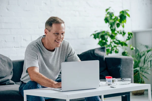 Bell'uomo in t-shirt sorridente e utilizzando il computer portatile in appartamento — Foto stock