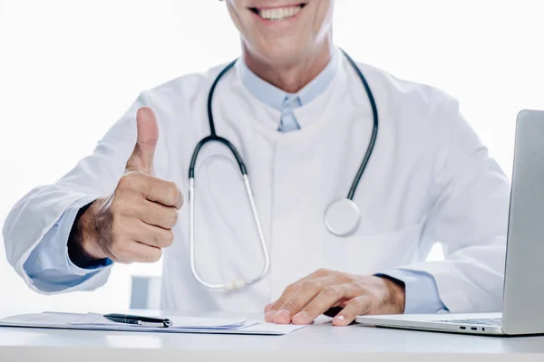 Vista recortada del médico en bata blanca sonriendo y mostrando el pulgar hacia arriba aislado en blanco - foto de stock