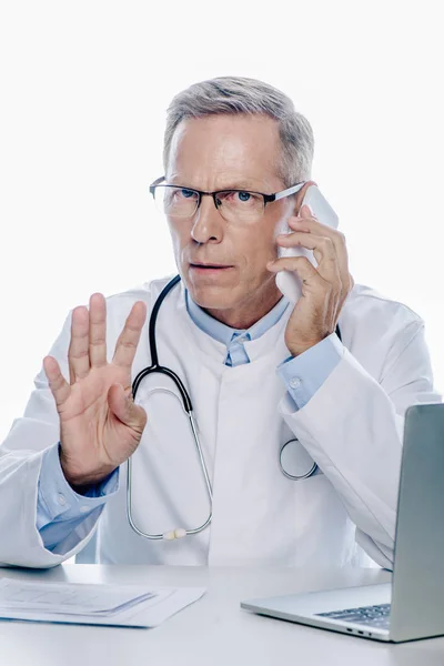 Beau médecin en manteau blanc parlant sur smartphone et montrant geste isolé sur blanc — Photo de stock