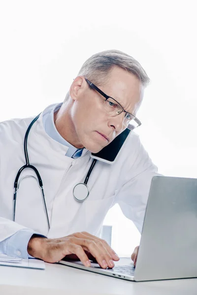 Schöner Arzt in weißem Mantel, der auf Smartphone spricht und Laptop isoliert auf weißem Grund benutzt — Stockfoto