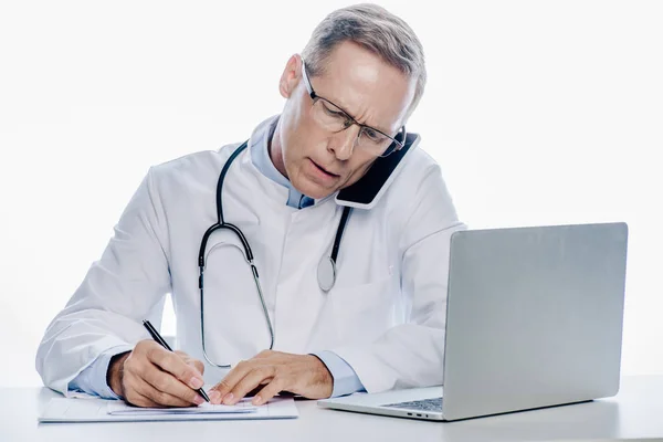 Handsome doctor in white coat talking on smartphone and writing isolated on white — Stock Photo