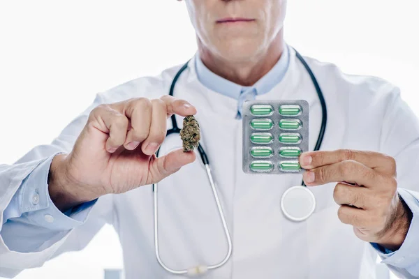Обрезанный взгляд врача в белом халате, держащего медицинскую марихуану и таблетки изолированы на белом — стоковое фото