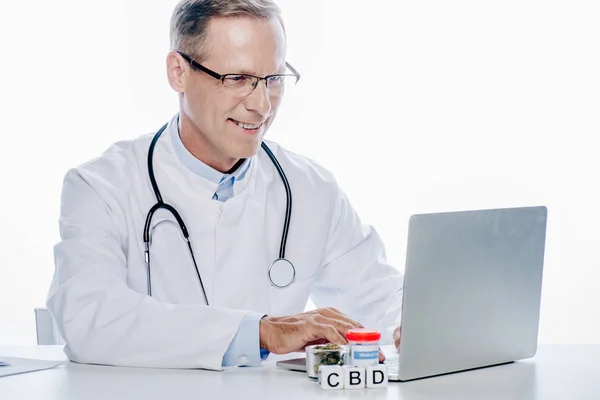 Schöner und lächelnder Arzt in weißem Mantel mit Laptop isoliert auf weiß — Stockfoto