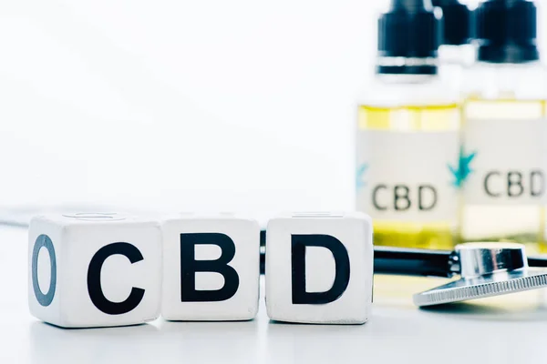 Messa a fuoco selettiva dell'olio di cannabis in bottiglie con lettere cbd, cubi e stetoscopio su sfondo bianco — Foto stock