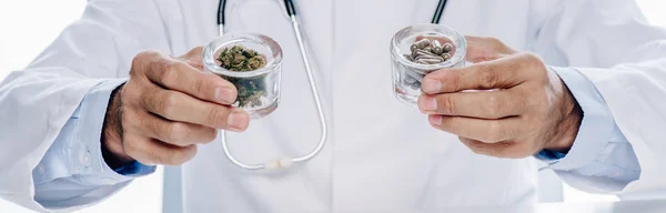 Inyección panorámica de médico de bata blanca sosteniendo píldoras y cannabis medicinal aislado en blanco - foto de stock