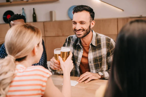 Sonriente hombre tintineo vasos de cerveza ligera con mujer joven mientras está sentado en el pub con amigos multiculturales - foto de stock