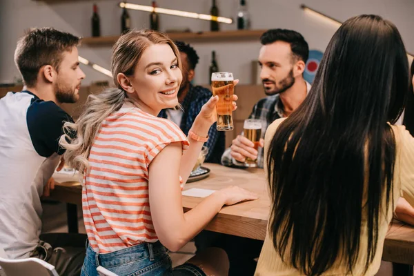 Messa a fuoco selettiva di allegra giovane donna sorridente alla fotocamera mentre tiene in mano un bicchiere di birra leggera — Foto stock