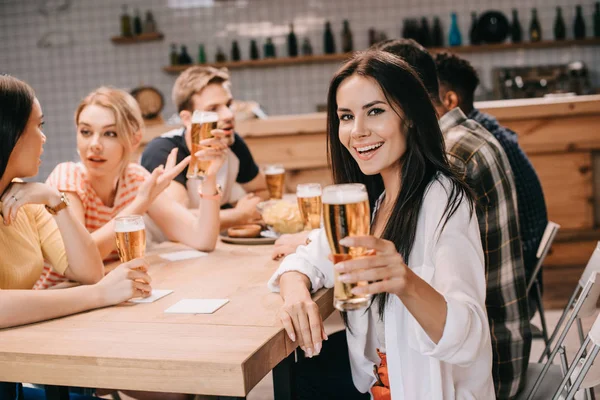 Alegre joven mujer sosteniendo un vaso de cerveza ligera y mirando a la cámara mientras se sienta cerca de amigos en el pub - foto de stock