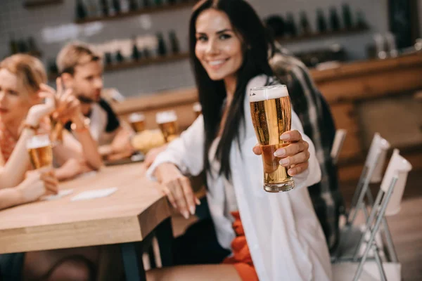 Selective focus of cheerful young woman holding glass of light beer and looking at camera while sitting near friends in bar — Stock Photo