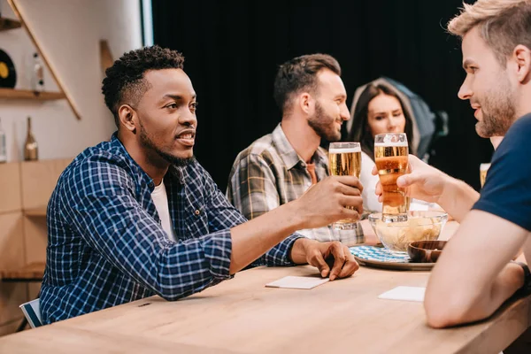 Amigos multiculturales tintineando vasos de cerveza ligera mientras pasan tiempo juntos en el pub - foto de stock