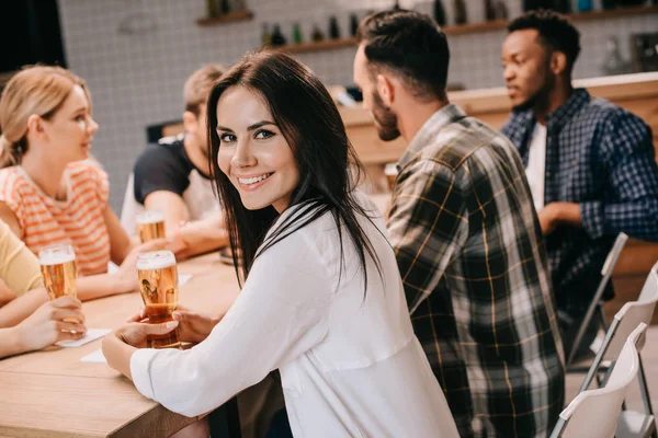 Attraktive junge Frau lächelt in die Kamera, während sie mit multikulturellen Freunden zusammen in der Kneipe sitzt — Stockfoto