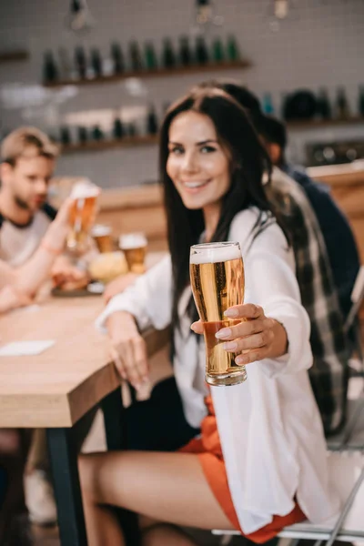 Messa a fuoco selettiva della giovane donna che tiene un bicchiere di birra e sorride alla macchina fotografica mentre è seduta in un pub insieme agli amici — Foto stock