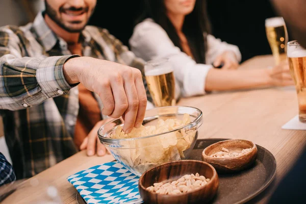 Abgeschnittene Ansicht eines Mannes, der Chips aus der Schüssel nimmt, während er mit Freunden in der Kneipe sitzt — Stockfoto