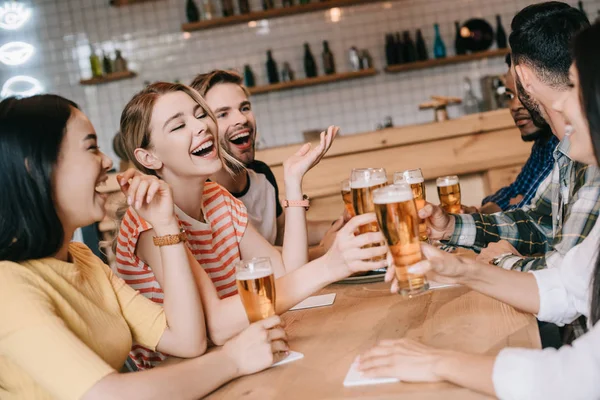 Alegres amigos multiculturales hablando y haciendo gestos mientras beben cerveza en el pub juntos - foto de stock