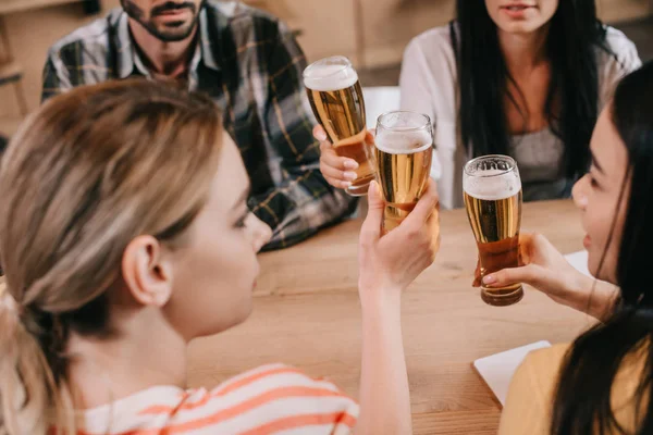 Vista recortada de amigos multiculturales tintineando vasos de cerveza ligera en el pub - foto de stock