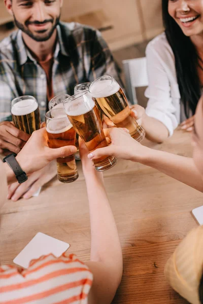 Vista parcial de amigos multiculturales tintineando vasos de cerveza ligera en el pub - foto de stock