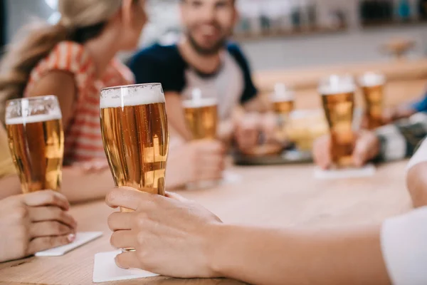 Vista recortada de las mujeres sosteniendo vasos de cerveza ligera mientras se sientan junto con amigos en el pub - foto de stock