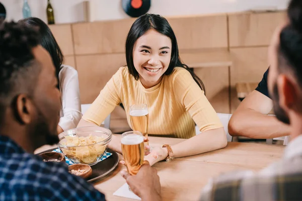 Selective focus of smiling asian girl holding glass of light beer while sitting in pub near multicultural friends — Stock Photo