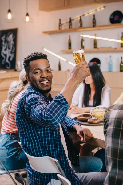 Alegre afroamericano hombre mirando a la cámara mientras sostiene un vaso de cerveza cerca de amigos multiculturales - foto de stock