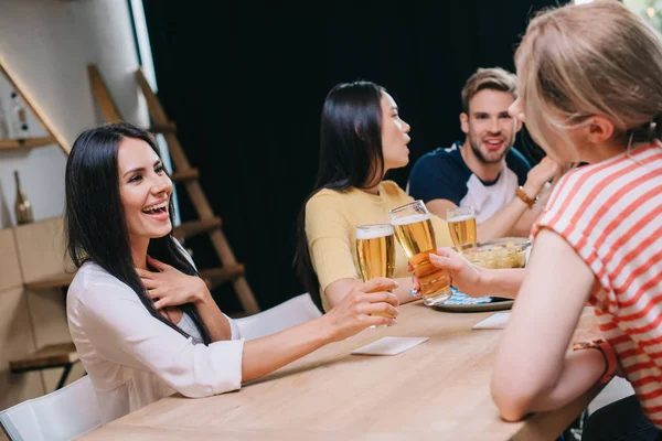 Alegre joven mujer tintineo vasos de cerveza ligera mientras se sienta en el pub con amigos multiculturales - foto de stock