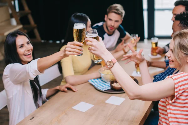Happy young woman clinking glasses of light beer while sitting in pub with multicultural friends — Stock Photo