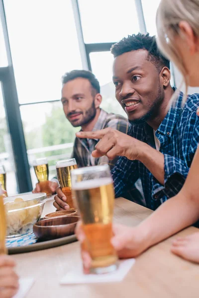 Messa a fuoco selettiva di allegro uomo afroamericano che punta con il dito mentre si siede con amici multiculturali nel pub — Foto stock