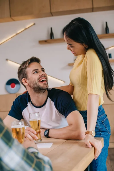 Joven asiático mujer de pie cerca amigo sentado con vaso de cerveza en pub - foto de stock