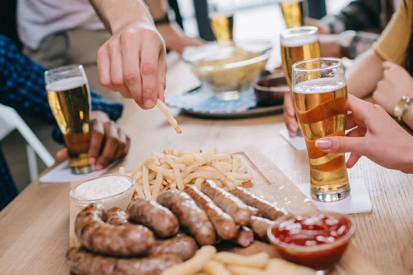 Cropped view of man taking french fries and fried sausages on wooden tray in pub — Stock Photo